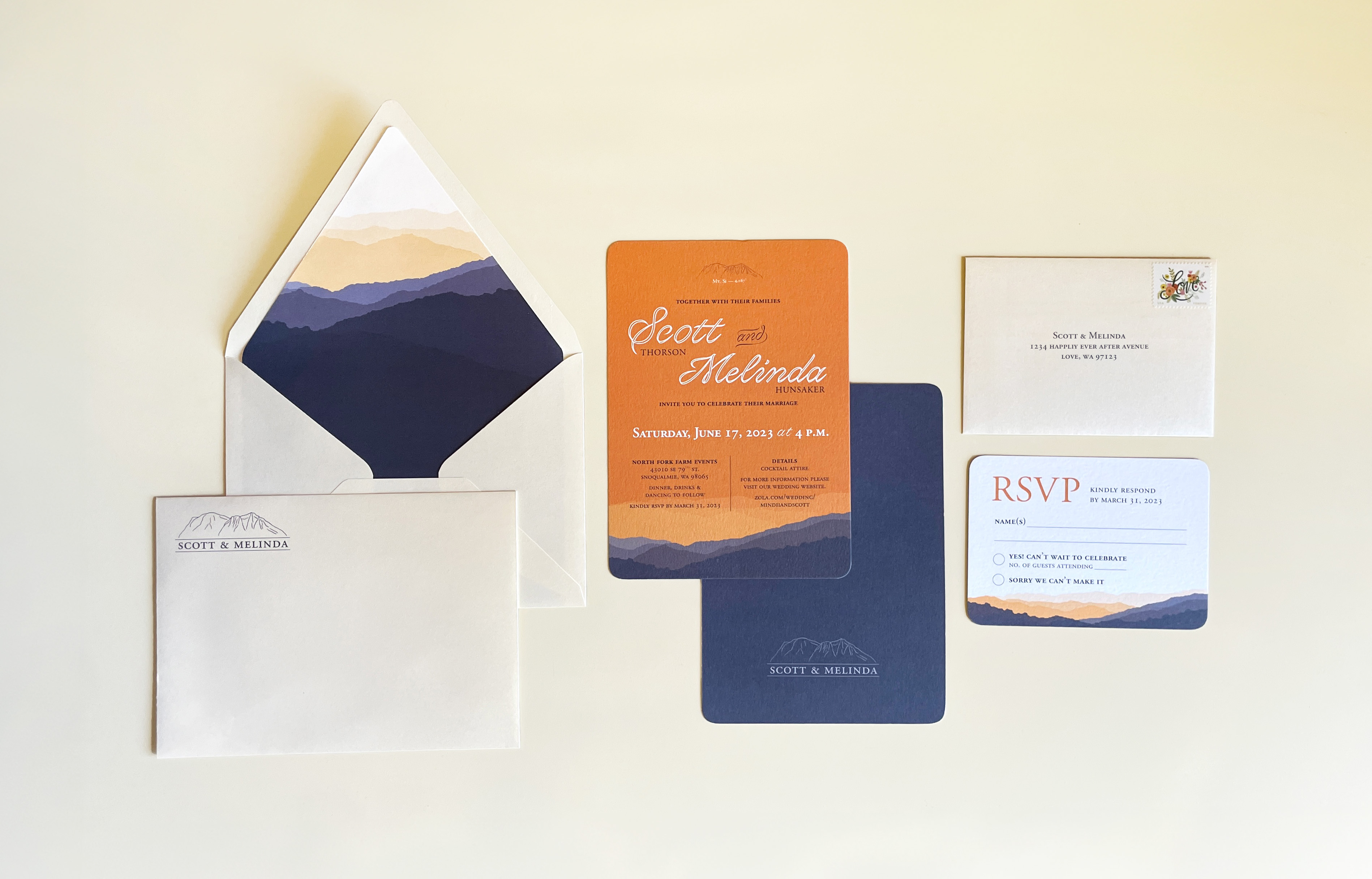 Flatlay of the entire wedding invitation package, including a champagne envelope, a contrasting sunset orange envelope sleeve with a navy mountain scape, the flat card invite, and the reply device.